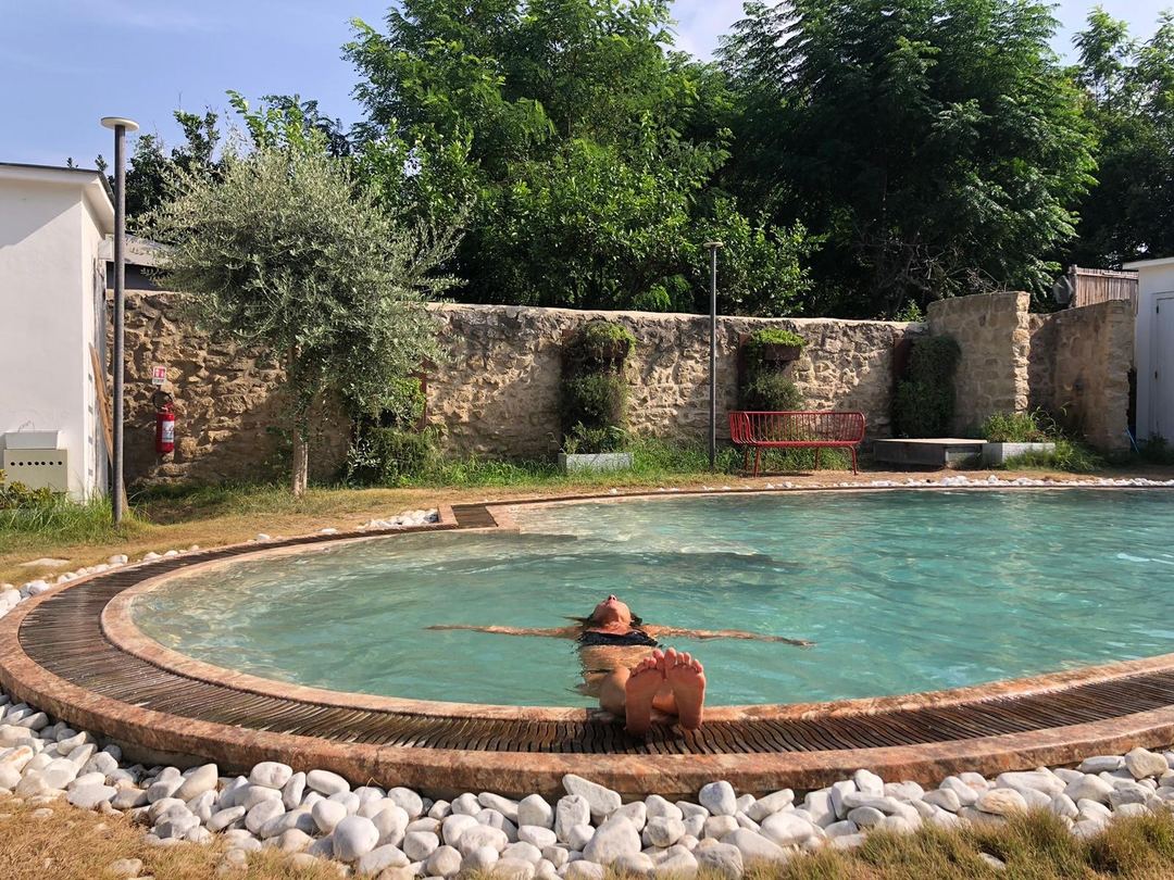 A swimmer floats in a circular thermal pool surrounded by wooden decking and white stones, with rustic stone walls and lush vegetation in the background.