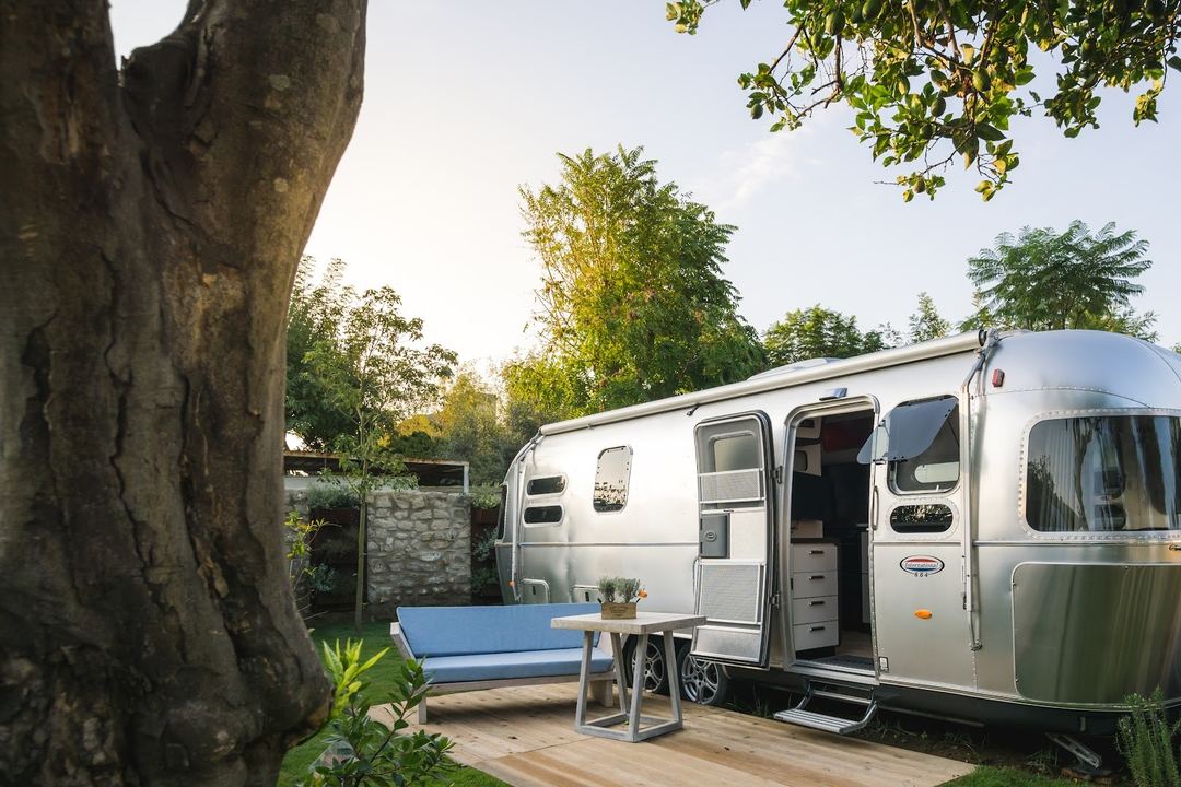 A silver Airstream travel trailer parked on a wooden deck beside a large tree, with a blue bench and small table in the foreground, surrounded by lush green vegetation.
