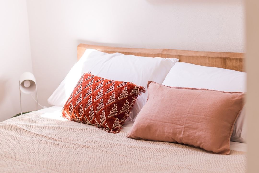 A neatly arranged bedroom bed with white pillows, a decorative red and gold patterned cushion, and a peachy-pink throw pillow against a wooden headboard.